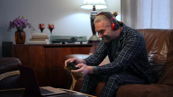 Engrossed Adult Man in Headphones Gaming with Joystick Online Sitting on Couch in Living Room alt