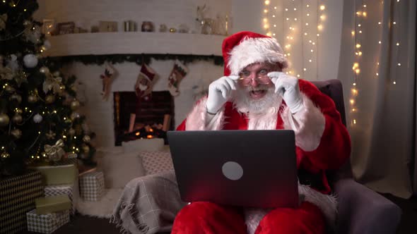 Modern Santa Claus. Cheerful Santa Claus Working on Laptop and Smiling While Sitting at His Chair