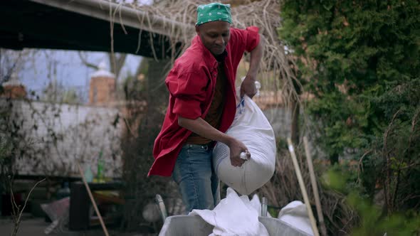 Young Strong African American Man Putting Heavy Bag with Building Substance in Construction Trolley alt