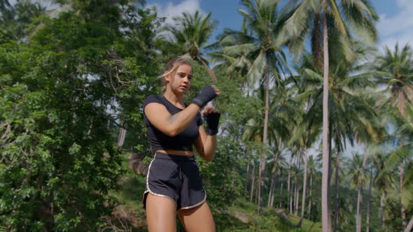 Sporty Girl in Black Shorts Practicing Boxing with Hands in Fingerless Gloves Among Tropical Palms alt