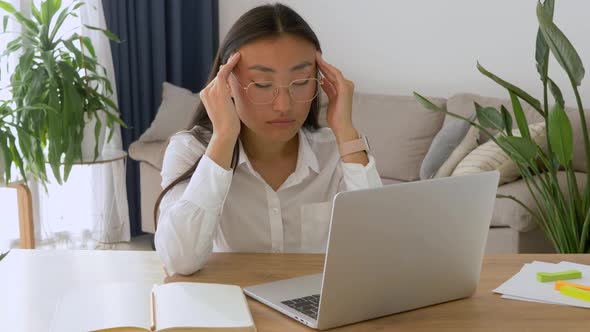 asian woman working at office desk in front of laptop suffering from chronic daily headaches alt