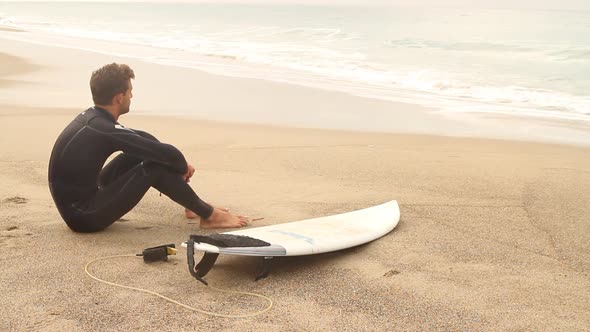 A young male surfer sitting on the beach looking out towards the ocean. alt