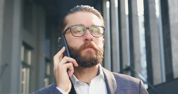 Bearded Businessman in Glasses which Talking on Mobile with Business Partner Outdoors alt