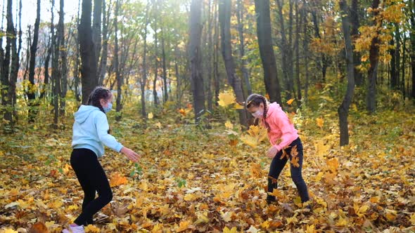 Two girls in medical masks throw fallen yellow leaves in the Park on an autumn day
