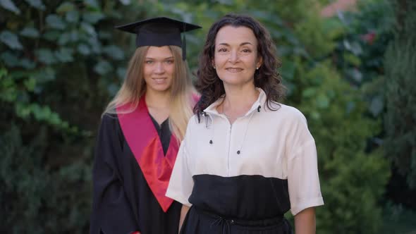 Portrait of Mature Happy Woman Posing with Blurred Graduate in Gown and Mortarboard at Background alt