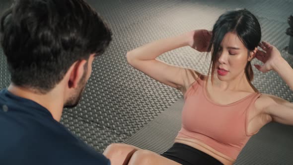 Asian young beautiful sport woman training sit up in gym with trainer holding legs in fitness club. alt