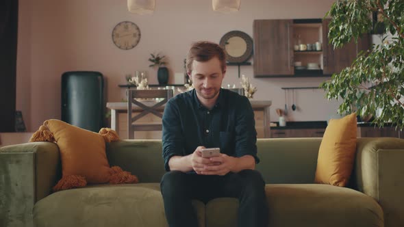 A Young Man is Sitting on the Sofa at Home Using a Smartphone