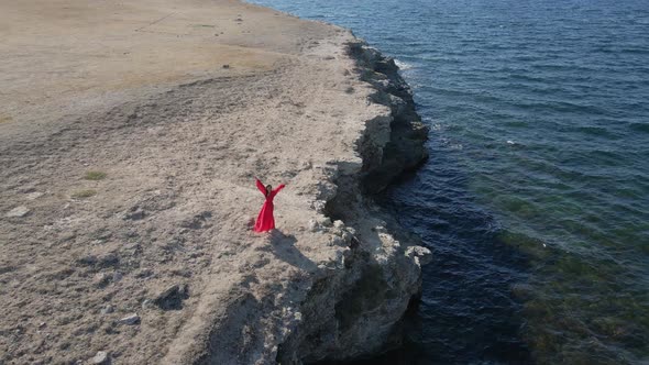 Brunette Woman in a Red Long Dress Stands on the Edge of a Cliff By the Sea alt