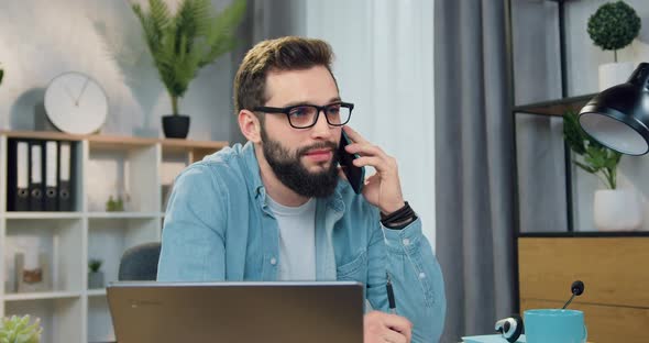 Guy in Glasses Talking on Mobile Sitting in front of Computer in His Home Office alt