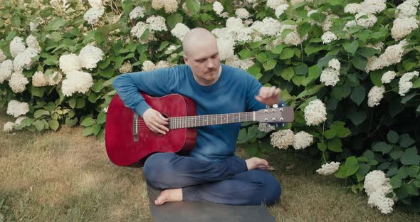 Man with an Acoustic Guitar is Sitting in a Park in Nature and Tuning Instrument