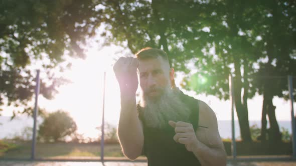 Middle Aged Man with Long Gray Beard Boxing with Shadow on Basketball Court During Sunrise Slow alt