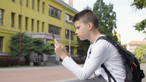 A Caucasian Teenage Boy Looks at a Tablet  Side View  a School in the Background