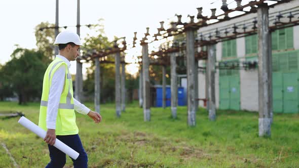 Construction Worker or Engineer Walking Near High Voltage Electrical Lines Towards Power Station