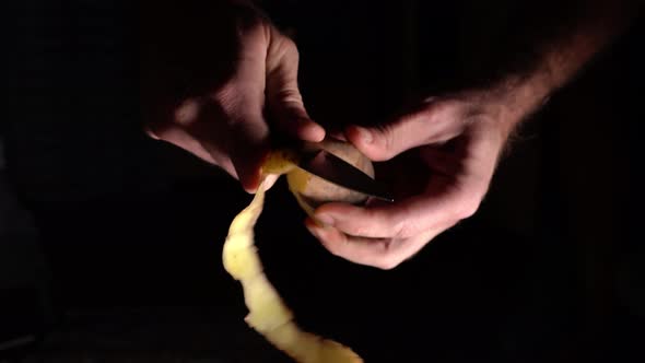 On A Black Background, A Man Peels A Potato With A Knife To Prepare Delicious Food