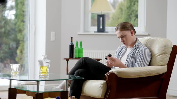 Wide Shot of Depressed Young Man with Gun Sitting on Armchair with Alcohol on Table alt