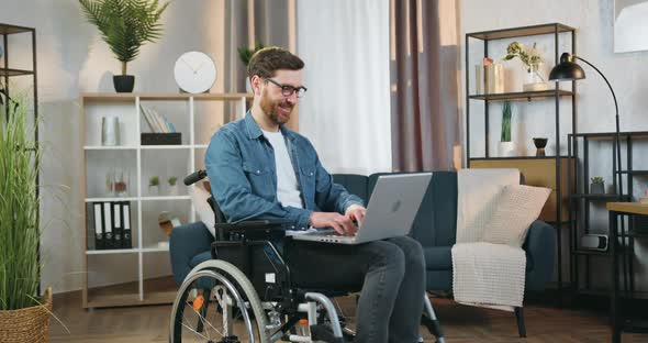 Man in Glasses Sitting in Wheelchair After Accident at Home and Working on Computer alt