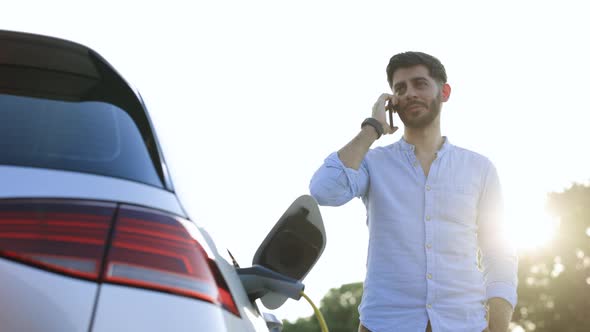 Joyful 30-aged Man With Beard Which Has Mobile Conversation While Luxurious Car Charging Battery
