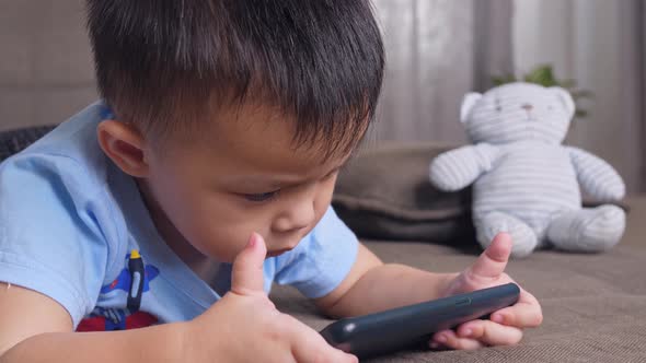 Asian Little Boy Laying On A Sofa Playing Smartphone alt