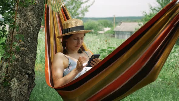 Portrait of young woman using mobile phone lying in hammock outdoors. alt