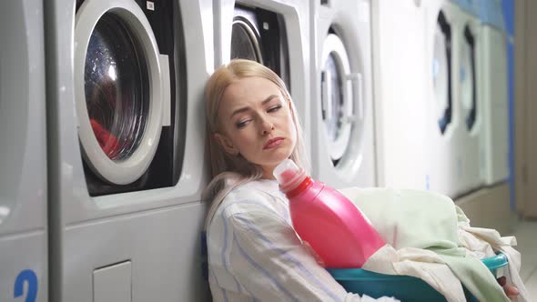 A Sad Young Blonde Sits at the Washing Machine in the Laundry Room and Waits for Her Underwear and