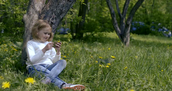 Cute Blonde Little Girl Is Sitting on Grass Leaning on Tree Trunk in Forest, Playing Smartphone alt