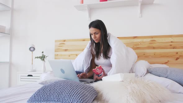 Сheerful Young Woman Using Laptop in Bed at Home Covered with Blanket and Eating Donut alt