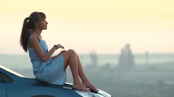 Happy Woman Driver in Blue Summer Dress Enjoying Warm Evening Laying on Her Car Hood alt
