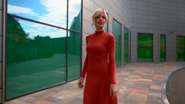 a Blonde Woman in a Red Dress and Black High-heeled Shoes Walks Along a Modern Building with Large alt