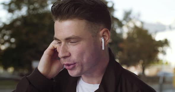 Portrait of Young Caucasian Student Cheerful Man with Wireless Headphone in Ear Having Phone Call alt