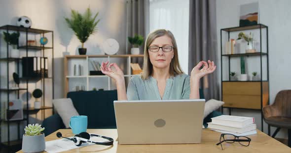 Senior Woman Meditating while Sitting at Table with Laptop alt