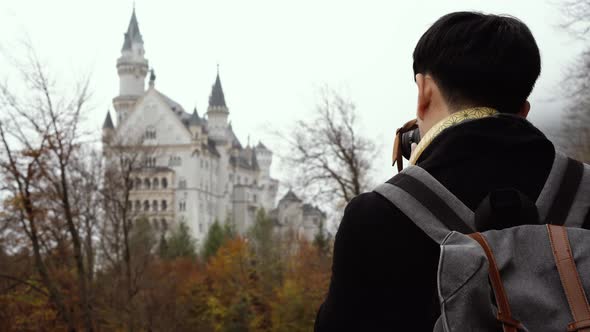 Male Asian Tourist with Backpack and Camera Standing and Enjoying View of Neuschwanstein Castle alt