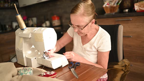 Confident Senior Elderly Woman Seamstress White Shirt and Glasses Sitting in Front of Sewing Machine