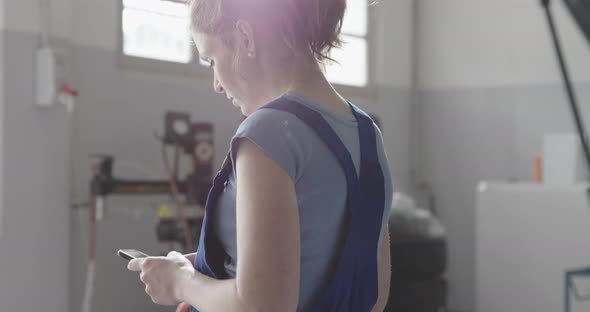 Professional female mechanic working in a car repair shop, she is messaging with a smartphone