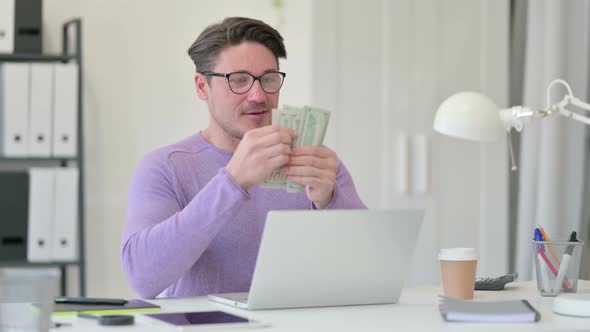 Middle Aged Man with Laptop Counting Dollar in Office  alt