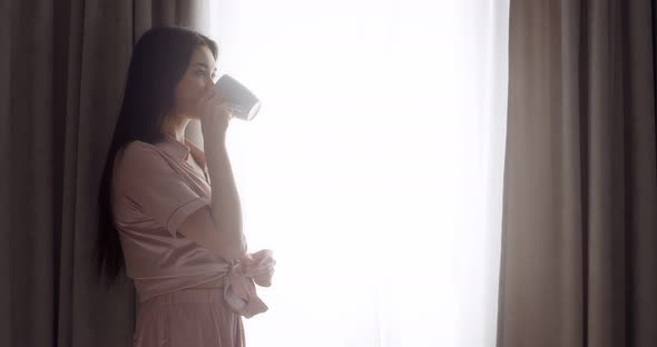 A woman stands at the window and drinks tea from a mug