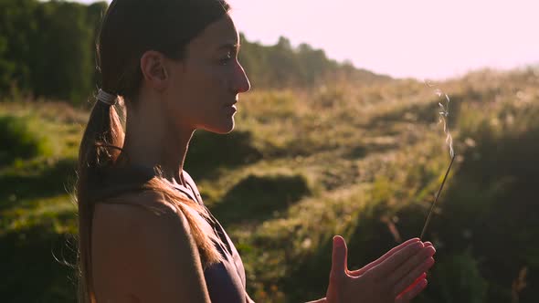 Woman Doing Yoga and Meditating in a Sunrise Outdoors, Holding Aroma Stick in Her Hands, Palms in