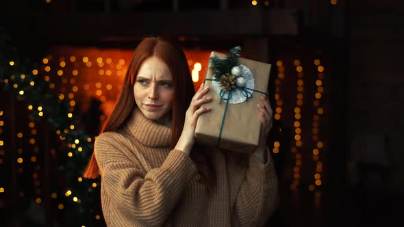 Portrait of Curious Beautiful Woman Shaking Gift Box with Christmas Presents Wrapped in Craft Paper. alt