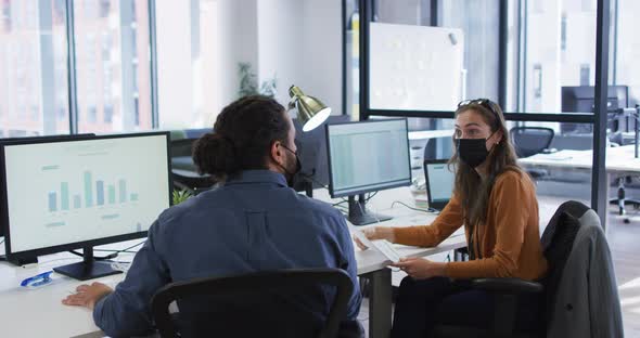 Diverse male and female colleague at desks using computers wearing face masks and elbow bumping alt