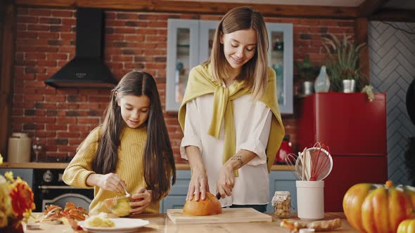 Little Daughter is Separating Pumpkin Seeds From Pulp with a Spoon and Her Smiling Mom is Cutting alt