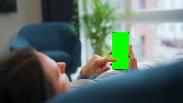 Woman Lies at Home on the Sofa and Using Smartphone with Green Mockup Screen in Vertical Mode alt