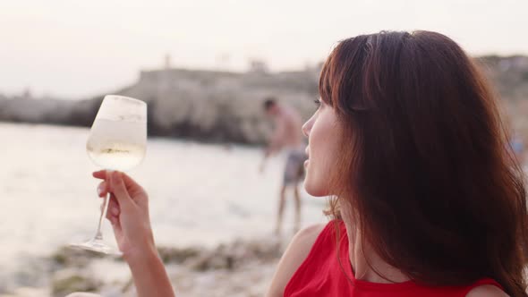 Young Brunette Woman in Red Shirt Sitting on Beach with Glass of Wine Enjoying Holidays