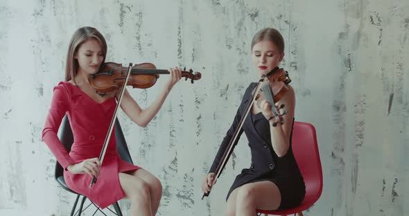 Two Elegant Girls in Dresses Playing the Violin Emotionally on Background Indoor alt