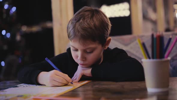 Cute Little Boy Drawing with Colour Pencils Sitting at the Table in Restaurant