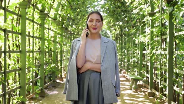 Young Woman Talking on the Phone in an Arched Corridor of Trees in the Garden alt