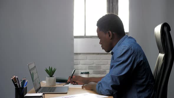African American Businessman Works for Laptop in Office Makes Notes in Notebook alt