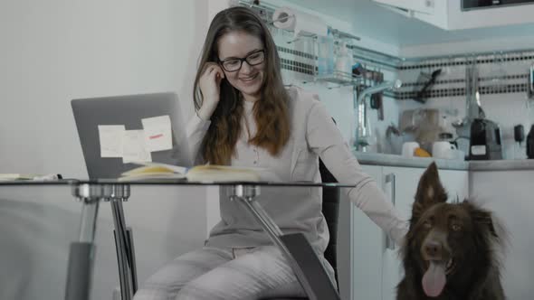 Young Woman At Home Sitting At The Table Using Her Laptop Close To Her Dog. alt