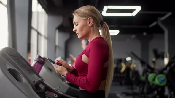Athletic Woman Does Cardio Training on Treadmill in Gym and Looks at Smartphone Side View alt