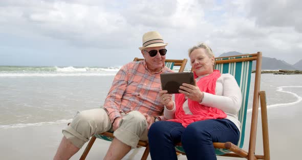 Front view of old caucasian senior couple using digital tablet and sitting on sunlounger a beach 4k alt