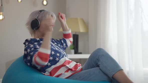 Side View Joyful Mature Woman in Headphones Dancing Sitting on Comfortable Bag Chair at Home in alt