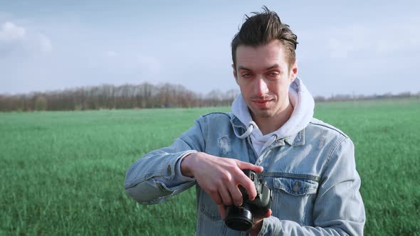 Young Male Photographer Taking Photo on Professional Photocamera Standing at Beautiful Green Field alt